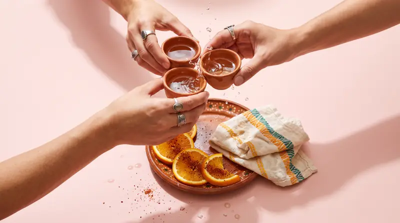 Three hands clinking clay copitas of mezcal over orange slices and sal de gusano