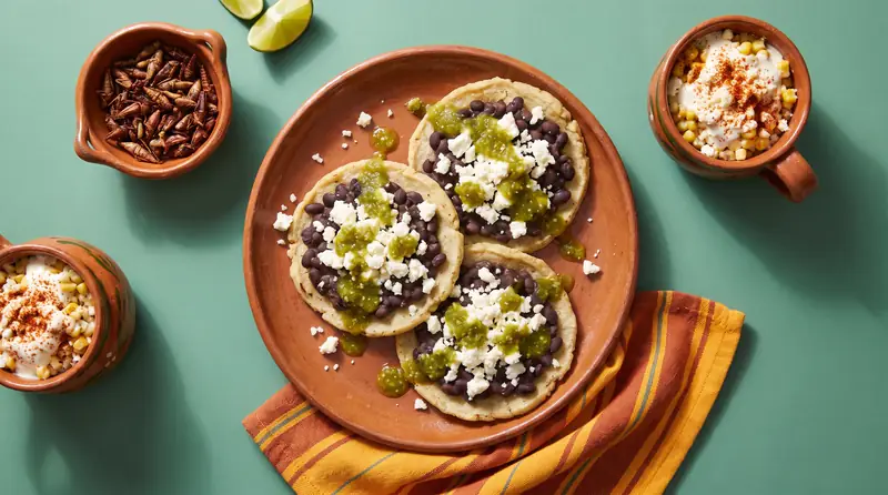 Three memelas with beans and queso fresco, a bowl of toasted chapulines with lime and a cup of esquites