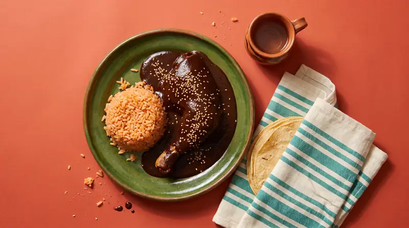Chicken under glossy black mole with sesame seeds, red rice and warm tortillas on a green clay plate