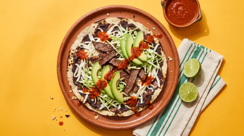 A wide crisp tlayuda topped with beans, quesillo, tasajo, avocado and cabbage on a clay plate